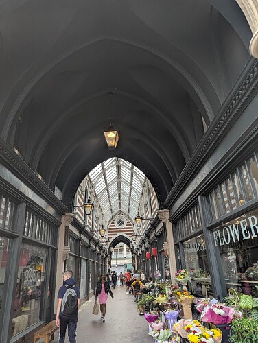 High quality image of an indoor walkway with shops. You can see a florist by the camera with people walking through.