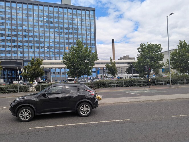 High quality image of part of a large hospital building. A bridge seems to connect two buildings together and you can see a pelican/pedex crossing as well as a black vehicle.