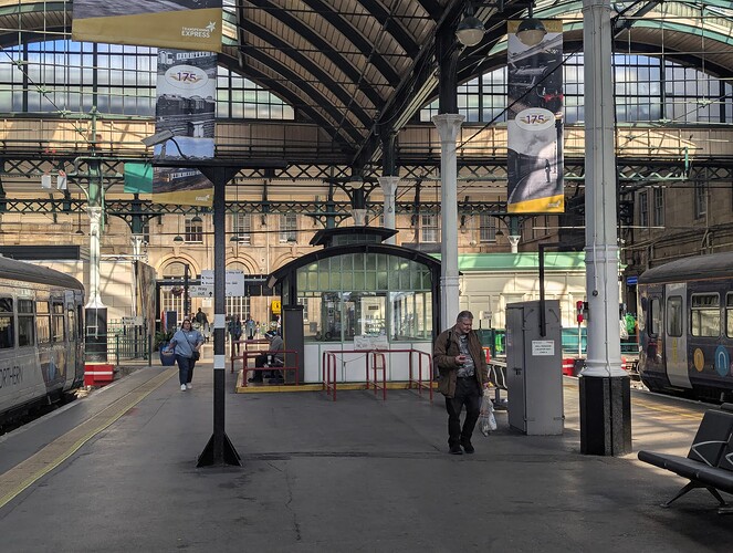 High quality image taken in an enclosed area of a railway station, Transpennine Express banners are hanging from the roof celebrating an anniversary. A Northern train can be seen on the left and right of the image and you can also see benches.