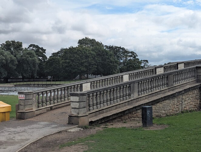 A high quality looking image of a bridge in a park, you can see a lake of some sort that it crosses over.