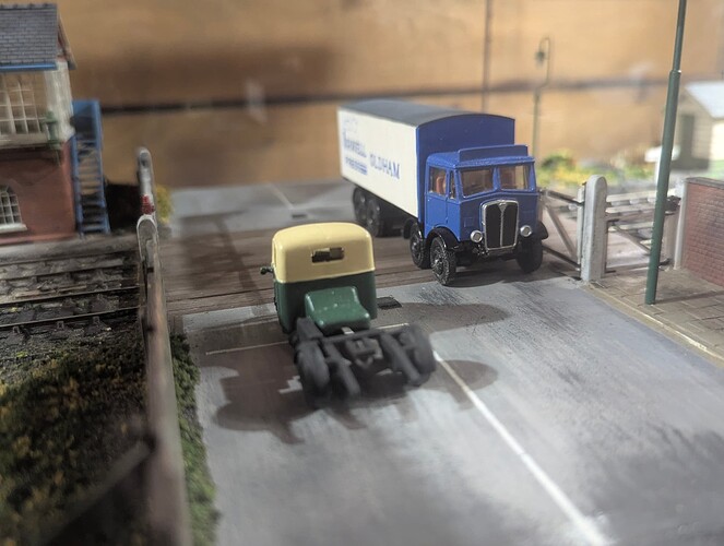 High quality image of a model railway crossing. A truck is currently on the crossing with another vehicle waiting to cross. A signal box can also be seen and reflections from the glass case that it was in.