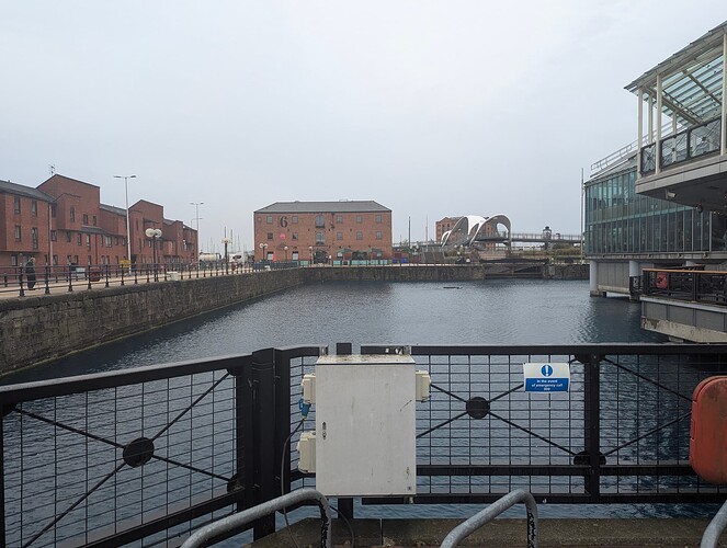High quality image from a bridge of some sort looking out to a shopping centre and another bridge further down. Various large brick buildings, cycle parking and part of what looks to be a lifebuoy can also be seen.