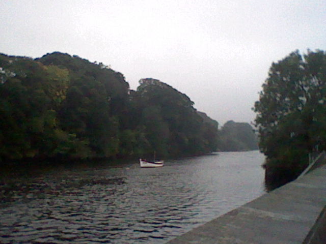 Low quality image of a boat going down a river, you can see parts of a wall and trees in the background